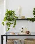 Glass console table with woven baskets, potted plants, and decor items against a white tiled wall.