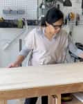Person working on a wooden board in a workshop with tools and a saw in the background.