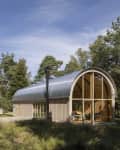 Curved-roof cabin with large windows surrounded by trees in a forest setting.
