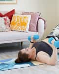 Woman exercising with blue dumbbells on a yoga mat, feet on a balance ball, in a living room with colorful pillows.