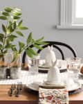Dining table with floral napkins, white ceramic decor, pastries, and green plant centerpiece.