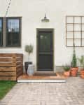 Black door on stucco wall with potted plants, wooden deck, and brick patio.