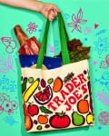 Reusable grocery bag with bread, greens, and packaged items, held by a hand against a colorful background.