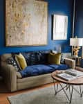 Living room with blue walls, beige sofa, abstract art, gold lamp, and round coffee table with books.