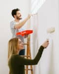 Two people painting a wall; one on a ladder with a roller tray, the other using a paint roller.