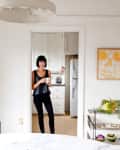 Woman in black top holding a mug, standing in a white kitchen doorway with a modern dining area.