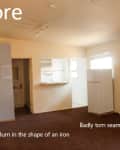 Empty room with brown carpet showing burn mark and torn seam, white walls, and a small kitchen area in the background.