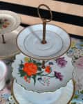 Two-tiered floral porcelain cake stand with gold trim on a floral tablecloth.
