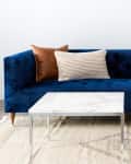 Blue velvet sofa with assorted pillows, a beige throw, marble coffee tables, and a lamp on a stack of books.