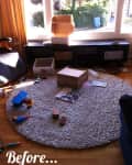 Messy living room with toys, boxes, and papers on a round rug near a window.