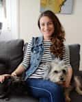 Woman in striped shirt and denim vest sitting on a gray sofa with two dogs.