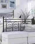 Black metal bed frame with striped bedding, white ottoman, wall art, and a vase with branches in a bright bedroom.