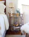 Bedside table with books, a lamp, a potted plant, and decorative items next to a white chair with a fur throw.