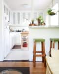 Open kitchen with white cabinets, wooden stools with green cushions, and hanging plants.