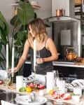 Woman setting a dining table with candles, pomegranates, and citrus fruits, next to large green plants in a modern kitchen.