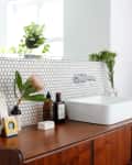 Bathroom vanity with hexagonal tile backsplash, white vessel sink, wooden cabinet, framed photo, and plants.