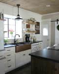 Farmhouse kitchen with white cabinets, black countertops, wooden shelves, and a copper sink under a window with tulips.