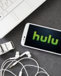 Smartphone displaying Hulu logo on desk with laptop, notepad, pen, earbuds, USB drive, and coffee cup.