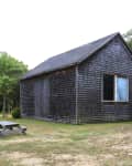 Wooden barn with shingle siding, large window, and sliding door, surrounded by grass and trees, with a picnic table nearby.