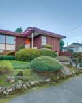 Mid-century modern house with large windows, red siding, and manicured shrubs in a suburban neighborhood.