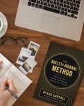 Hands writing in a journal on a wooden desk with a coffee cup, glasses, photos, and "The Bullet Journal Method" book.