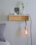 Wooden wall shelf with glass vases, photo frame, and hanging Edison bulb above a bed with gray pillows.