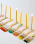 Paintbrushes with colorful bristles holding lit yellow candles in a row.