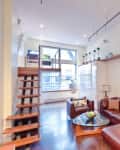 Loft-style living room with brown leather sectional, glass coffee table, wine fridge, and wooden staircase.