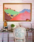 Eclectic desk setup with colorful abstract painting, geode, potted plant, and patterned chair against pink-striped wallpaper.