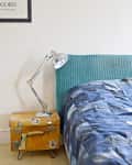 Bed with blue patchwork quilt, turquoise headboard, vintage suitcase nightstand, and silver desk lamp.