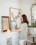 Woman arranging a large glass jar on a white sideboard, with a lit candle, framed art, and a decorative mirror nearby.