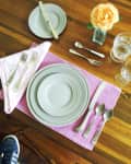 Table setting with gray plates, silver cutlery, pink placemat, and a yellow rose in a vase on a wooden table.