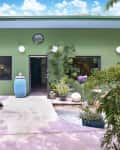 Green house exterior with large windows, potted plants, and a blue ceramic jar by the entrance.