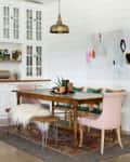Dining room with wooden table, pink and teal chairs, abstract wall art, large plant, and white cabinets with glass doors.