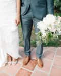 Bride in lace gown holding dress, groom in gray suit with brown shoes, holding white floral bouquet on terracotta tiles.