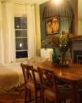Cozy dining room with wooden table, chairs, floral centerpiece, large mirror, and bed by curtained windows.