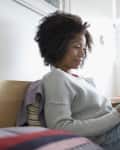 Woman in a gray sweater reading a book while sitting on a bed in a bright room.