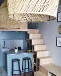 Small kitchen with blue cabinets, wooden stairs, two black stools, and woven pendant lights.