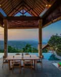 Open-air dining area with wooden table, chairs, and infinity pool overlooking a lush landscape at sunset.