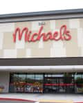 Entrance of Michaels store with large red sign, beige and gray facade, and potted plants by the doors.