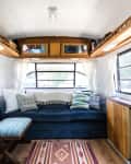 Renovated bus interior with blue sofa, patterned rug, wooden shelves, and large windows with white curtains.