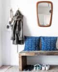 Two blue pillows on top of a simple wooden bench, potted plants are hanging from hooks