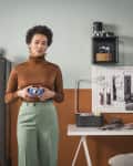 Woman in brown turtleneck holding a camera, standing in a room with vintage cameras and a modern desk setup.
