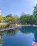 Curved swimming pool surrounded by stone path, lush greenery, and a two-story house.