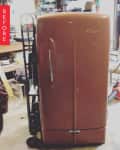 Vintage brown fridge on a dolly in a cluttered garage with tools and boxes.