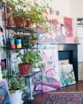 Art studio with floral paintings, potted plants, and an easel near a window, featuring a colorful rug and patterned cushions.