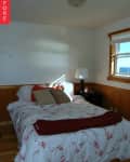 Bedroom with floral bedding, red throw, and pillows, wooden paneling, bedside lamp, and window with blinds.