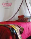 Dorm bed with red bedding, colorful pillows, and a DIY white canopy with tassels.
