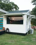 Modern tiny house with a blue door, wooden accents, and a small porch with chairs, set in a grassy area.