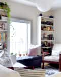 Cozy living room with bookshelves, a navy ottoman, white armchair, and potted plants by a window.